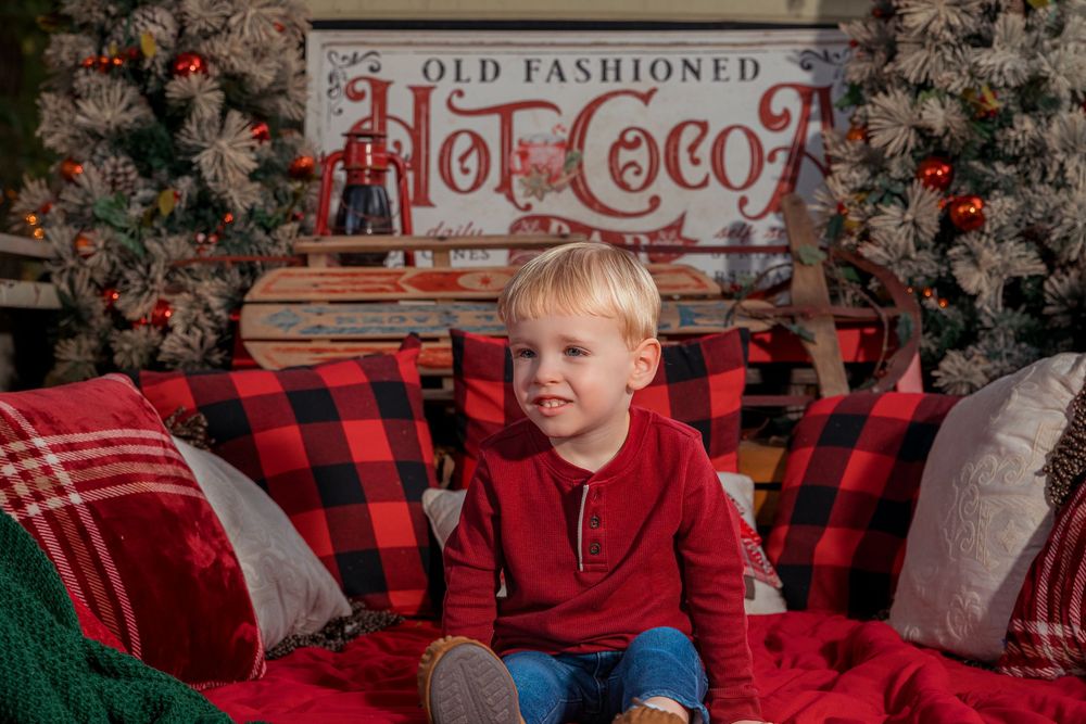 Holiday photo setup with buffalo plaid pillows and vintage hot cocoa sign against floral backdrop.
