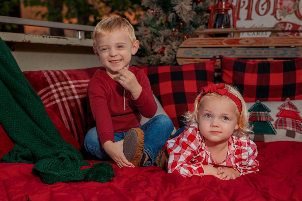 Two children pose on red plaid holiday decor with one wearing burgundy and one in buffalo check pattern.