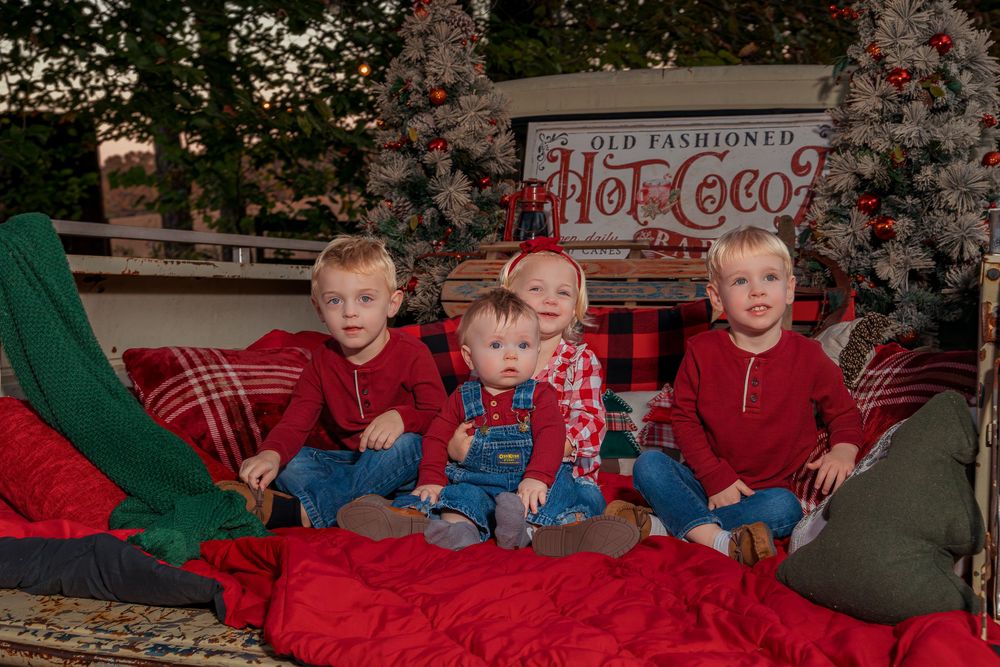 Three children in holiday attire sit on a red blanket in front of a Christmas tree for festive photos.