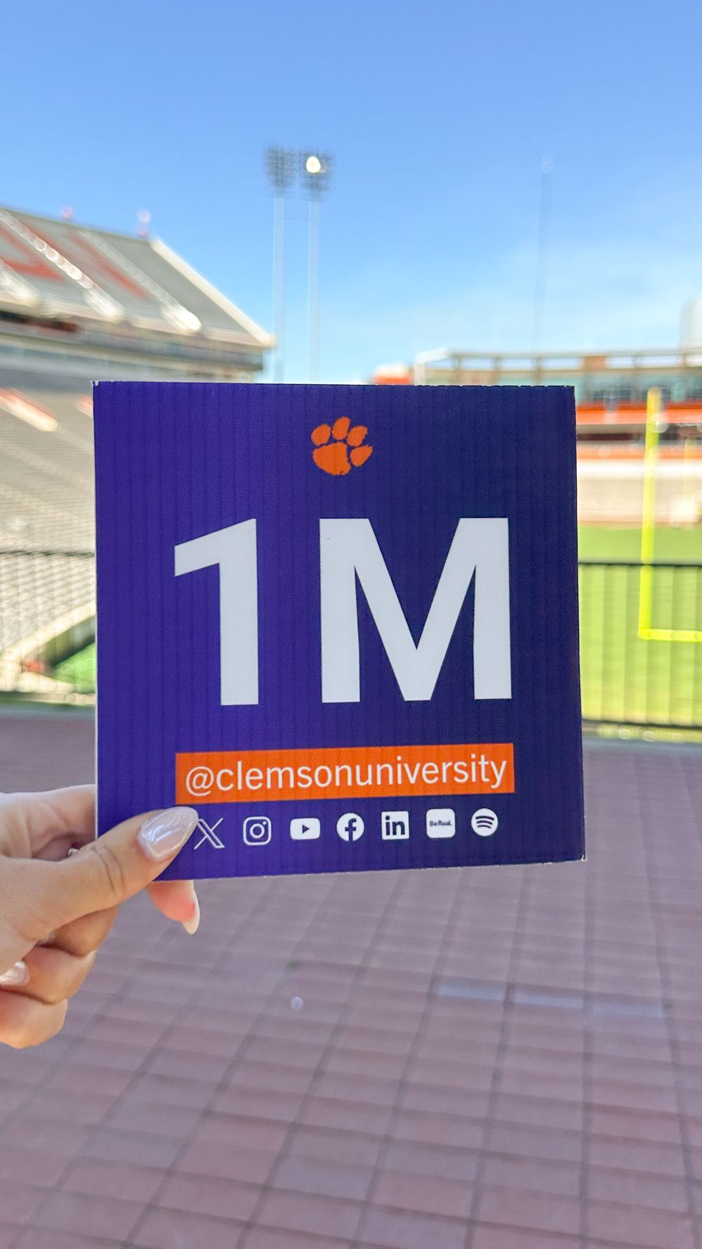 Purple social media milestone card showing 1M followers for Clemson University held up in stadium.