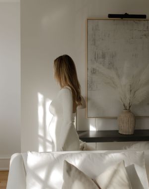 Silhouette of woman in white dress standing by window with sunlight casting dramatic shadows.