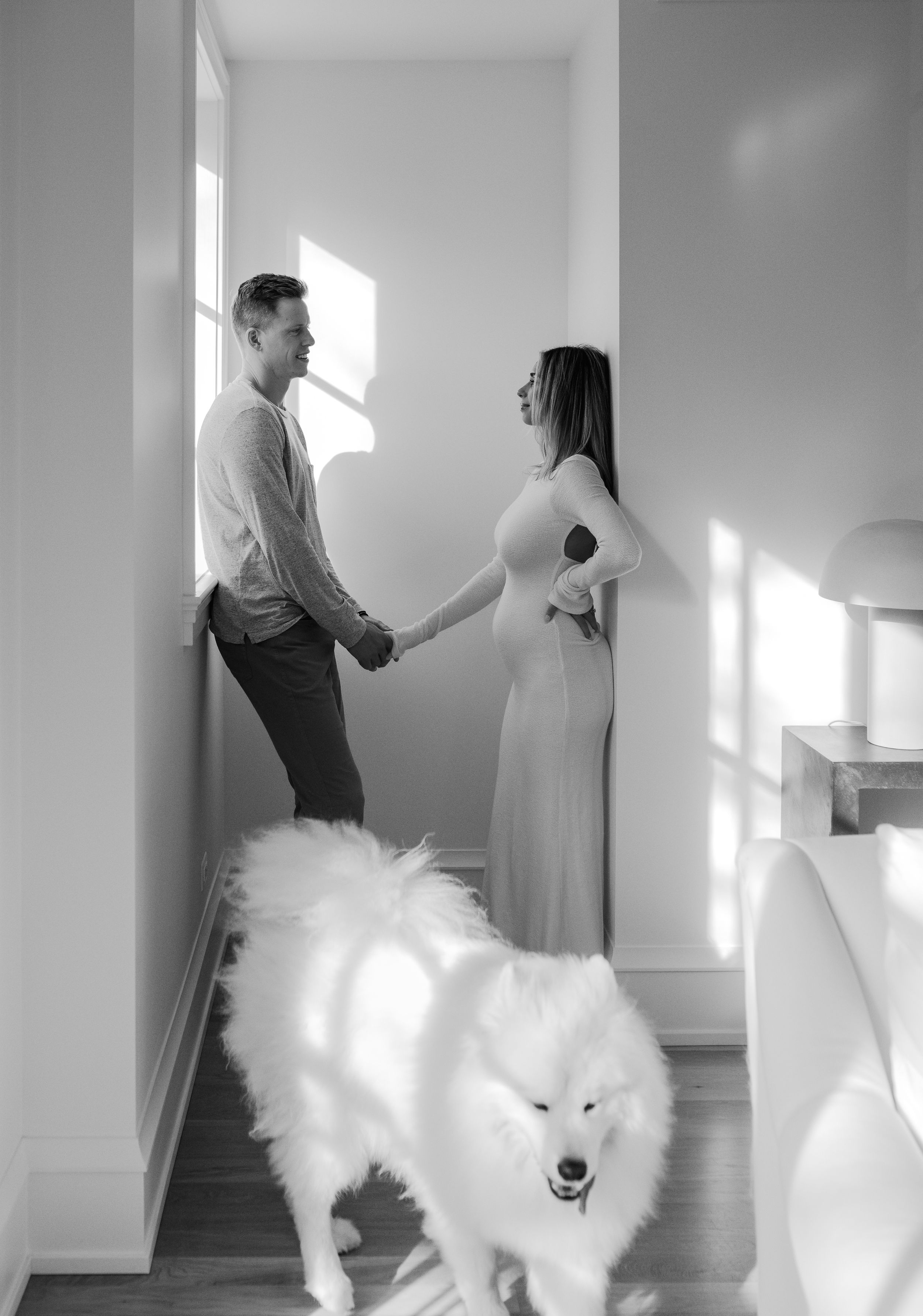 Black and white photo of couple holding hands in hallway with fluffy white dog in foreground.