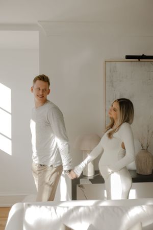 A couple shares a playful moment during their maternity photoshoot in a bright, modern living room.