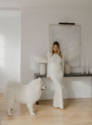 A glowing maternity photo featuring a white long dress and a fluffy white dog in a modern interior.