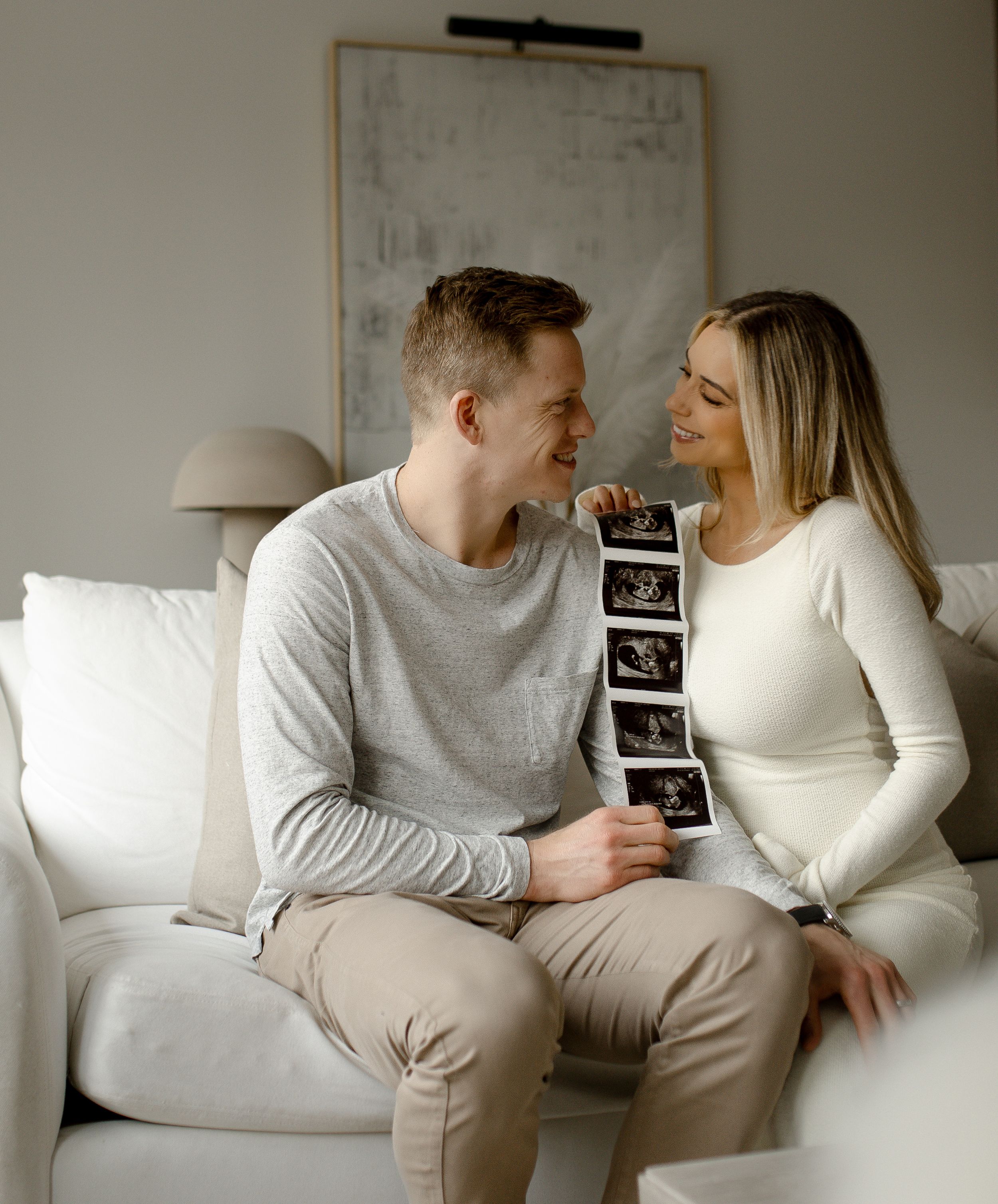 Couple sharing an intimate moment on white couch while holding ultrasound photos in a minimalist room setting.