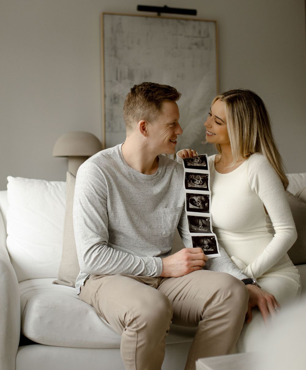Couple sharing an intimate moment on white couch while holding ultrasound photos in a minimalist room setting.