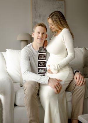 Expecting couple on white sofa sharing pregnancy announcement with ultrasound strip photos.