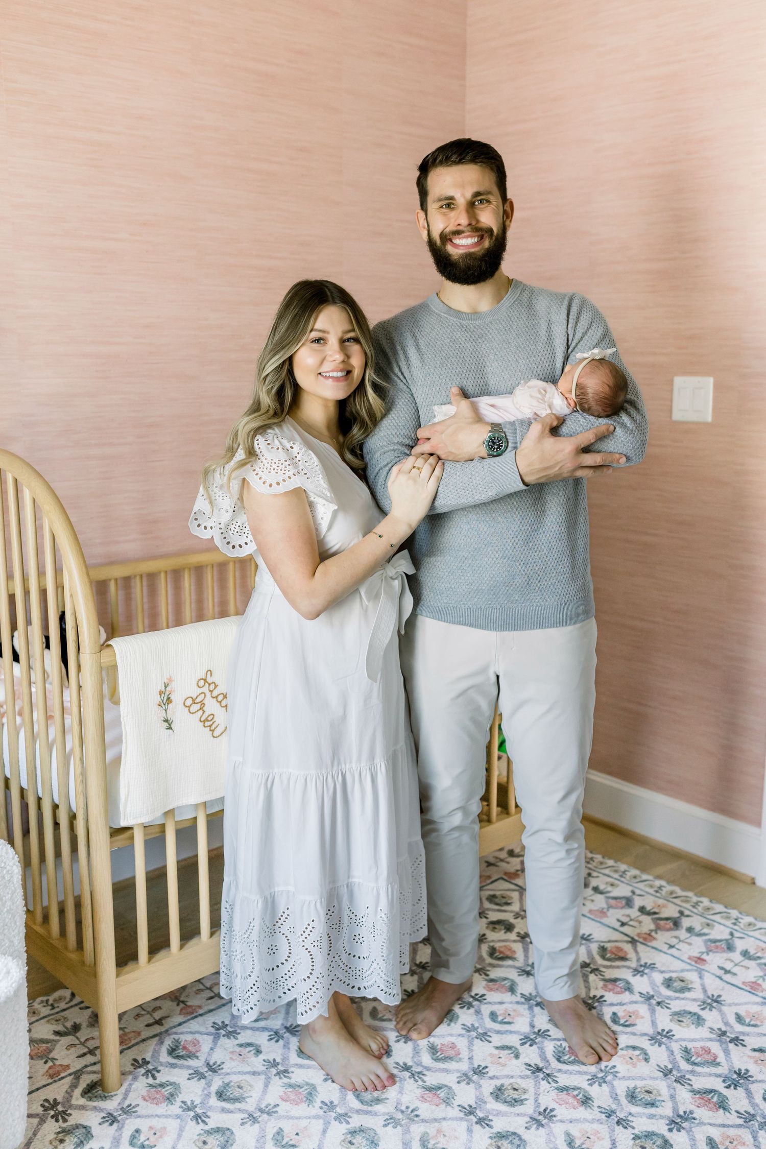 A couple in casual attire stands in a nursery holding their newborn baby near a wooden crib.