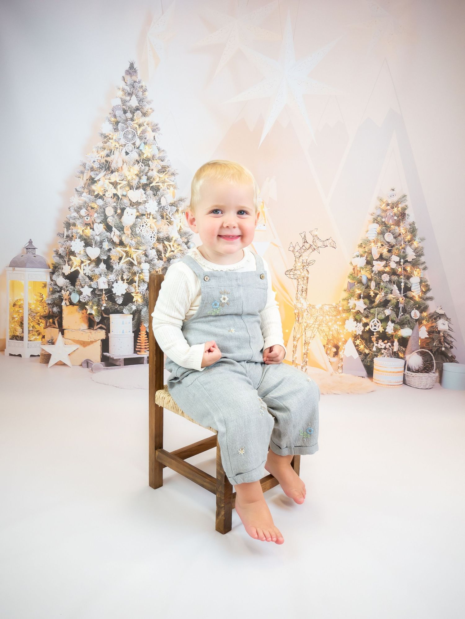 Small child sits on wooden chair in front of decorated white Christmas trees and glowing lanterns.