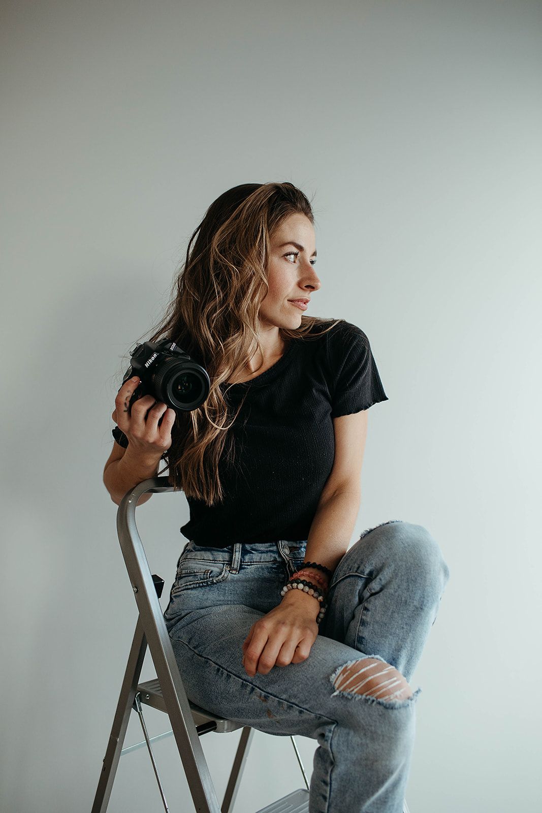 A photographer in casual attire sits on a folding chair holding a camera against a white backdrop.