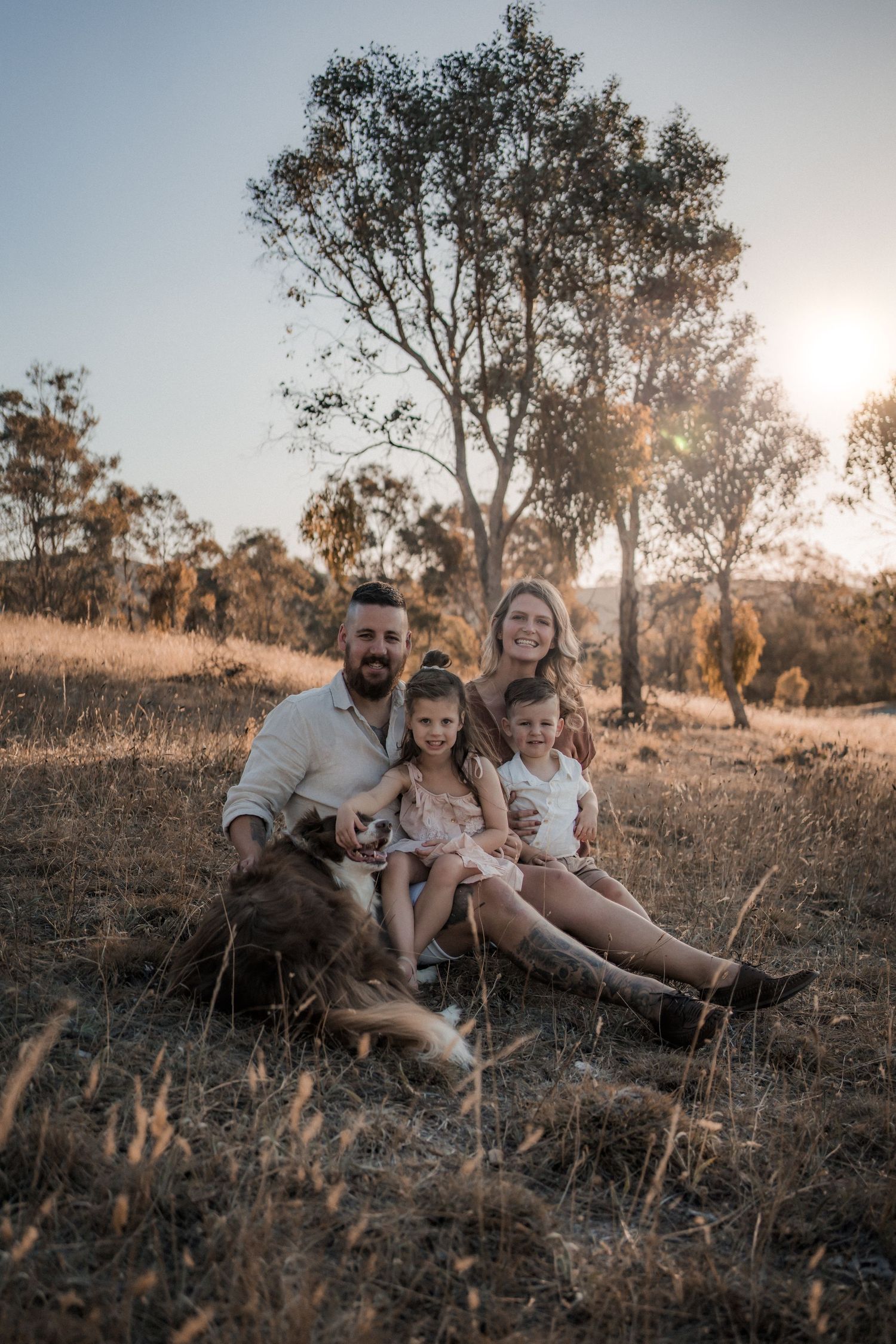 Family relaxing together on grassy hill during golden hour with tall trees in background.