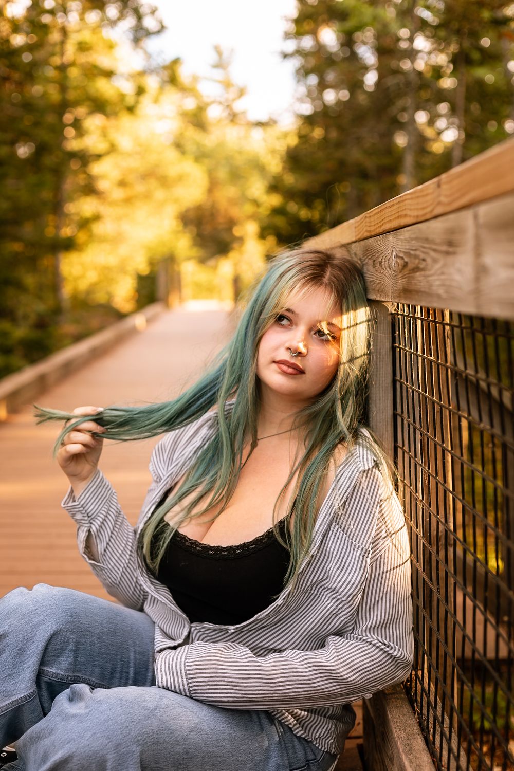 Young woman with teal hair leaning against wooden fence on bridge, wearing striped shirt, surrounded by autumn trees.
