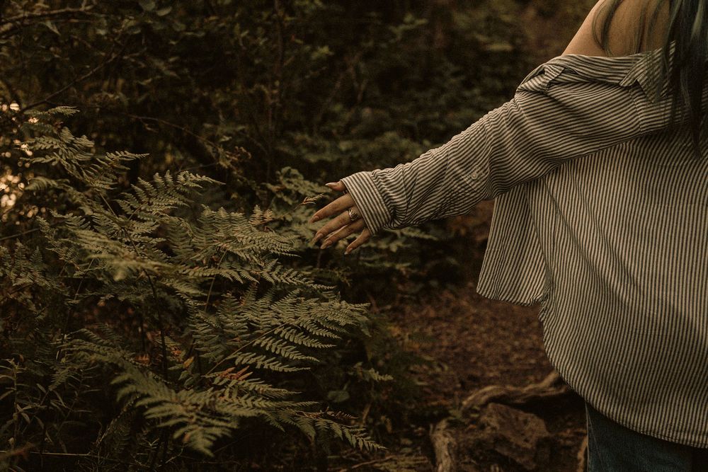 Pregnant woman in striped top gently touching green fern leaves in a lush forest setting.