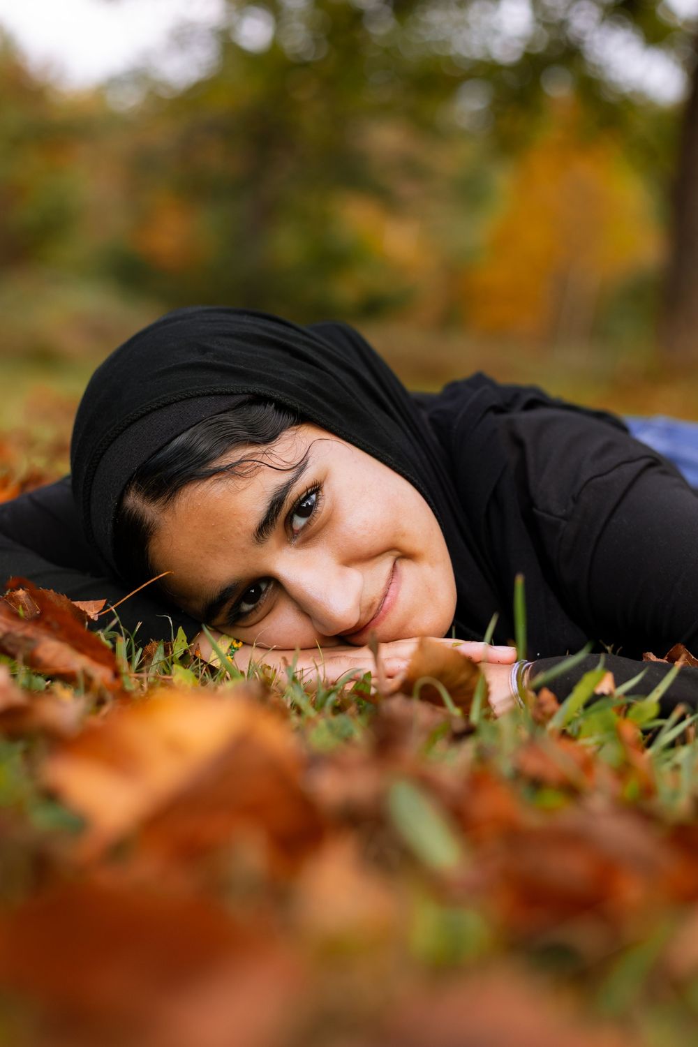 Person wearing black hijab lying in autumn leaves with a gentle smile.