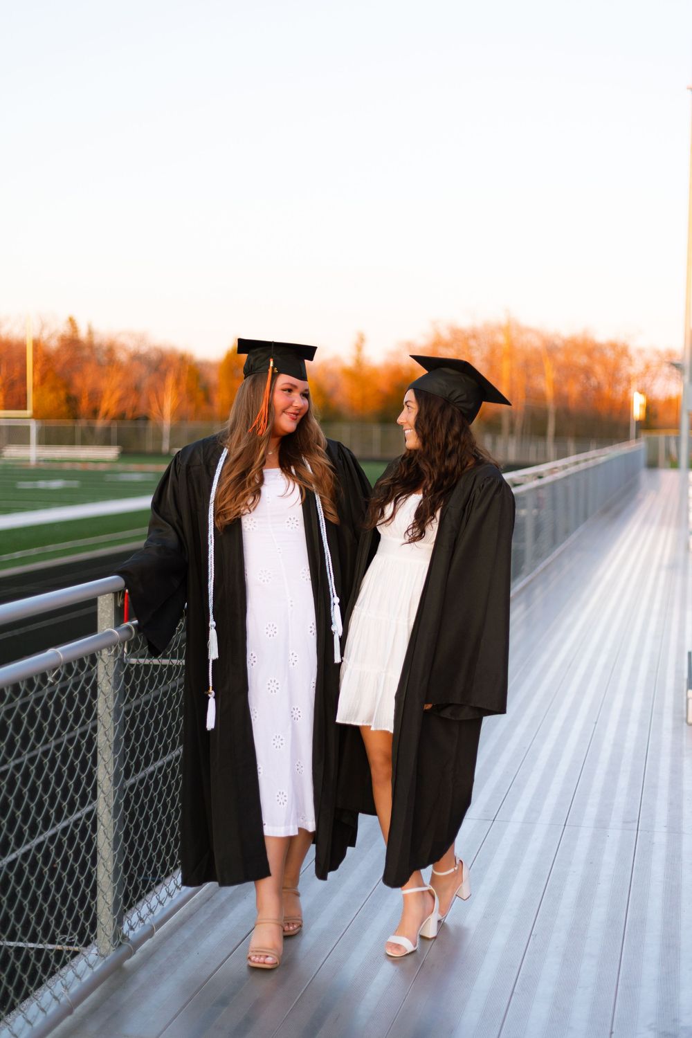 Two female graduates in black caps and gowns over white dresses smile at each other on stadium bleachers at sunset.