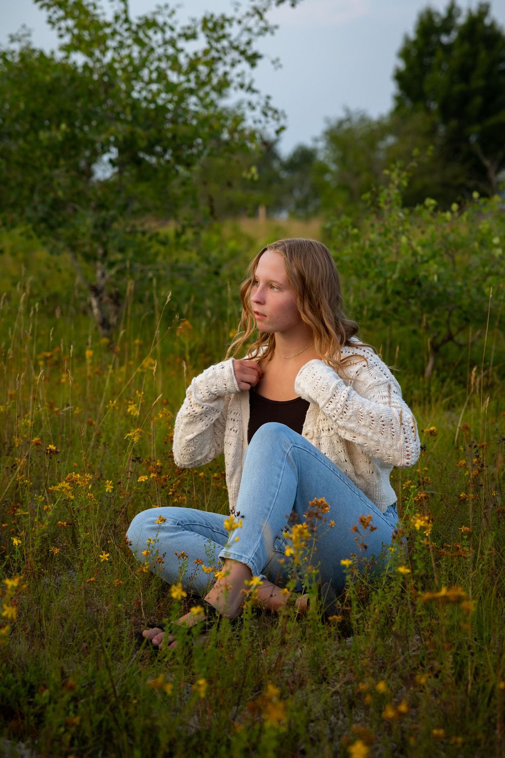 A person in a white sweater sits thoughtfully among wildflowers in a summer meadow.