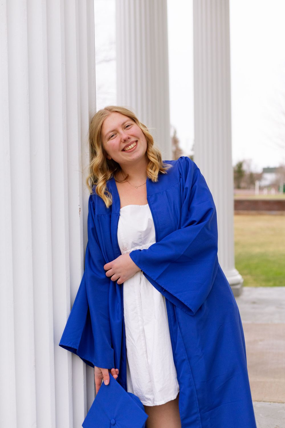 Smiling young woman in blue graduation gown and white dress leaning against white columns outdoors.