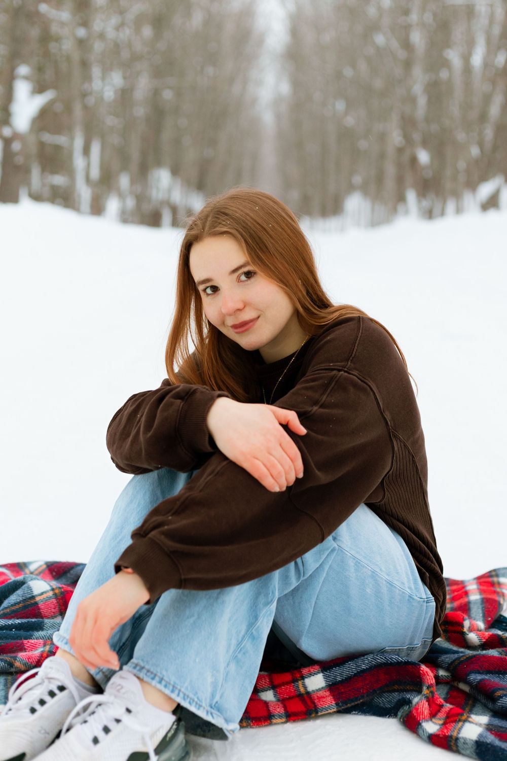 Young woman in brown sweater and jeans sits on plaid blanket in snowy winter landscape, smiling at camera.