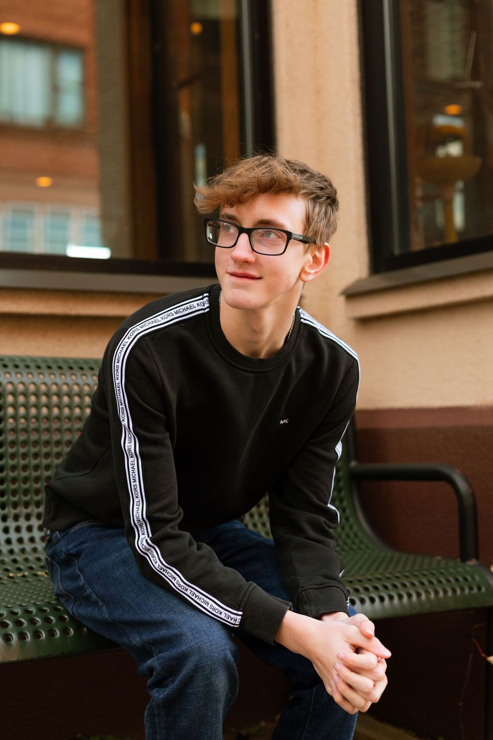 Person in glasses and black sweater with white stripes sitting casually on bench in outdoor urban setting.