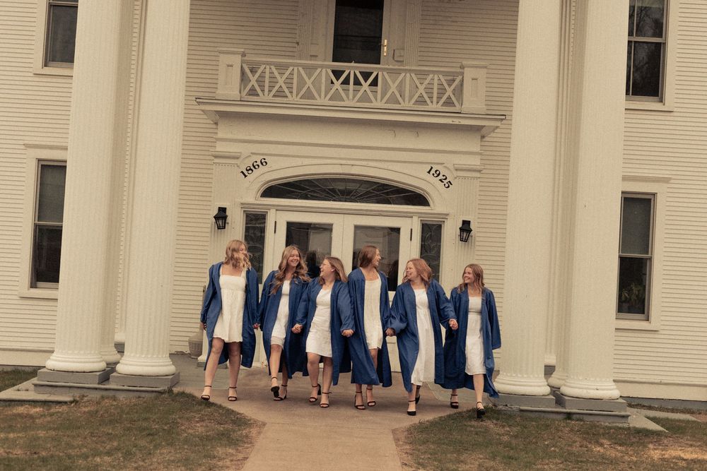 Group of female graduates in blue caps and gowns walking in front of a white colonial building.