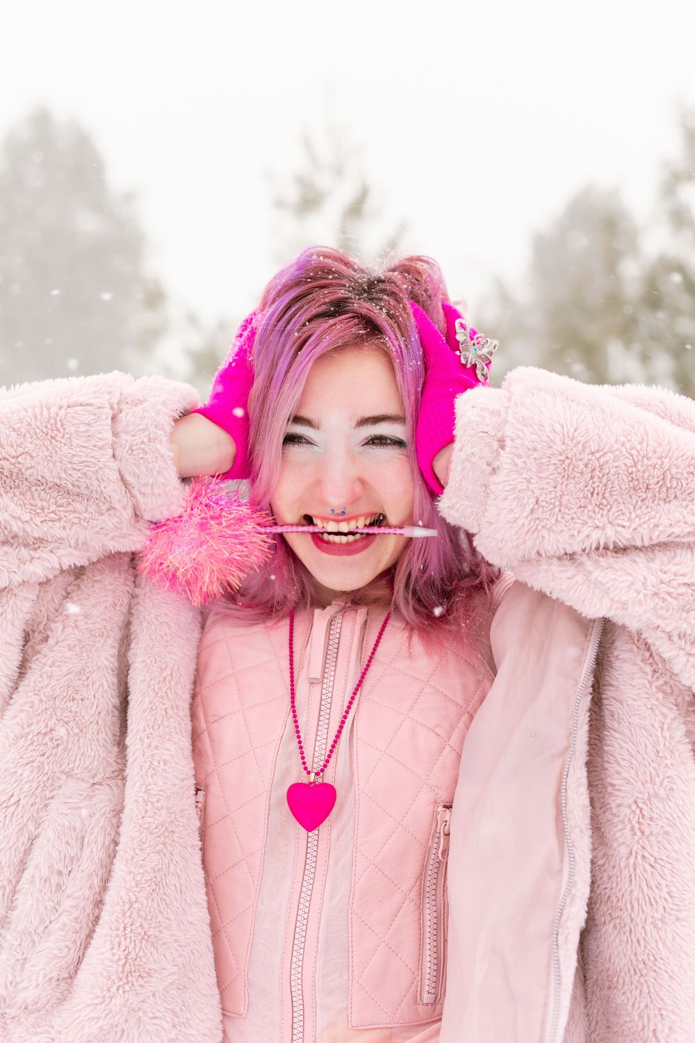 Young woman with pink hair in pink fuzzy coat and mittens poses playfully in snowy winter setting.