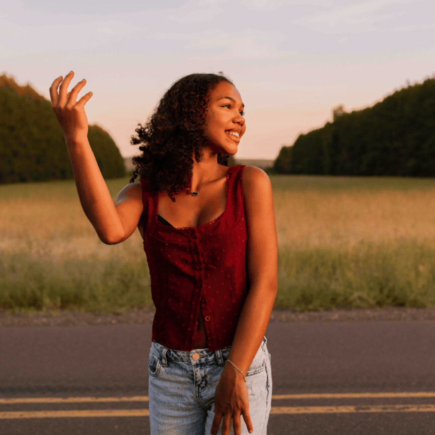Young woman in red top and jeans waving outdoors at sunset on a road surrounded by greenery.
