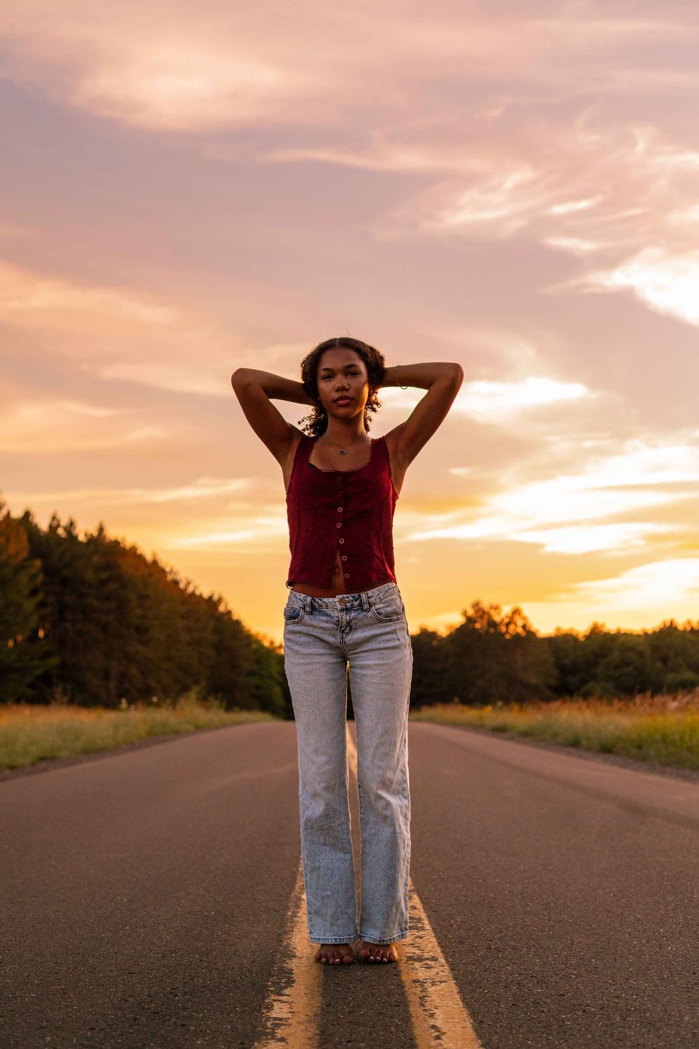 Young woman in red top and wide-leg jeans poses on empty road at golden sunset with trees in background.