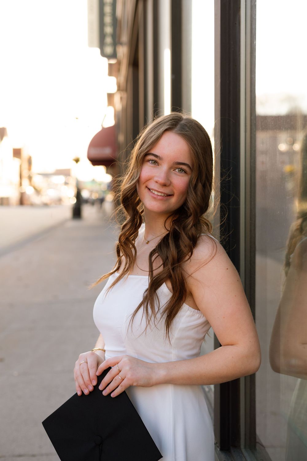 Young woman in white dress holding black clutch, smiling on urban sidewalk at golden hour.