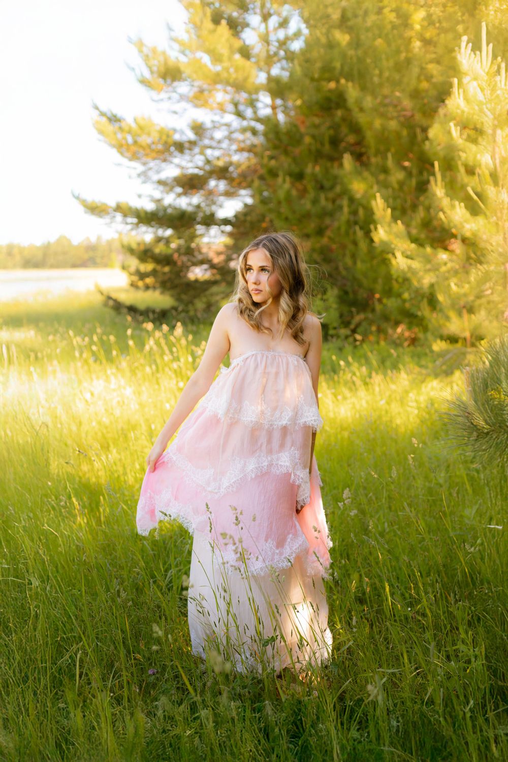 Young woman in flowing pink dress standing in sunlit green meadow with tall trees in background.