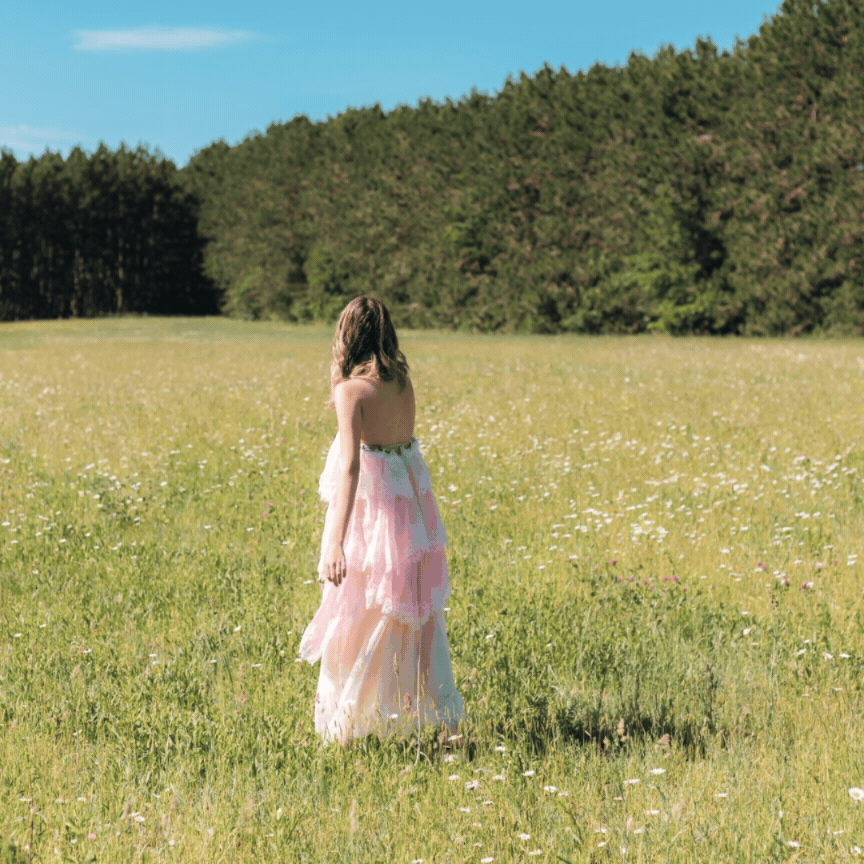 A woman in a flowing pink gown stands in a sunny green meadow with wildflowers and a dense tree line behind her.