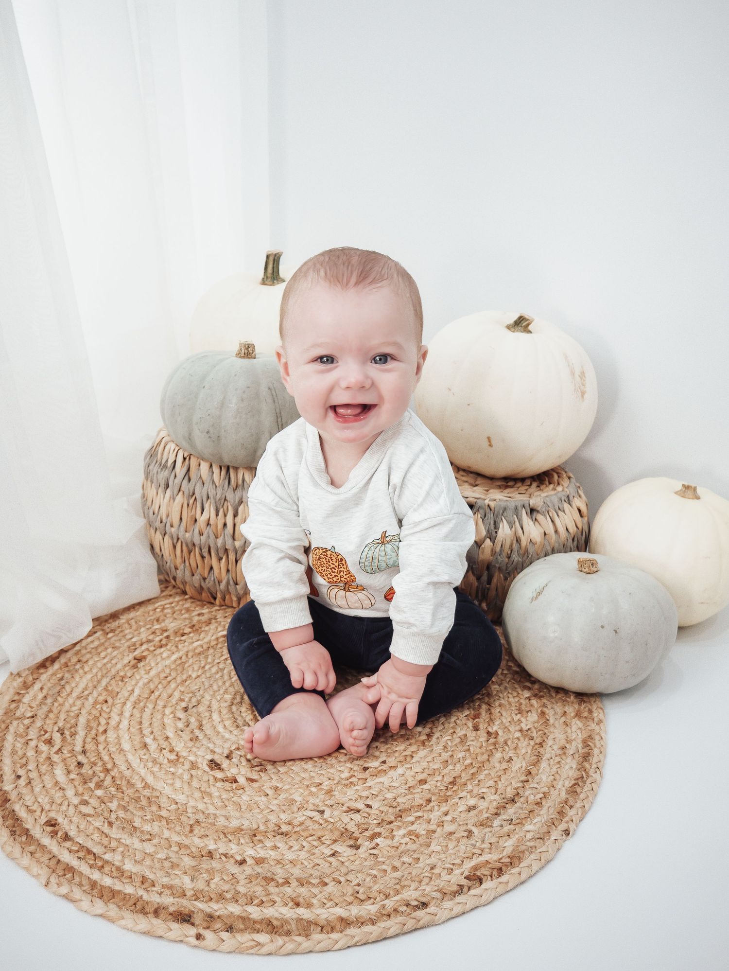 Smiling baby in white sweater sits on woven mat surrounded by white pumpkins.