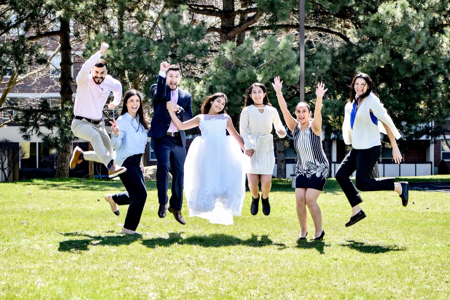 Joyful wedding party jumping in the air on green lawn surrounded by tall pine trees. Toronto GTA