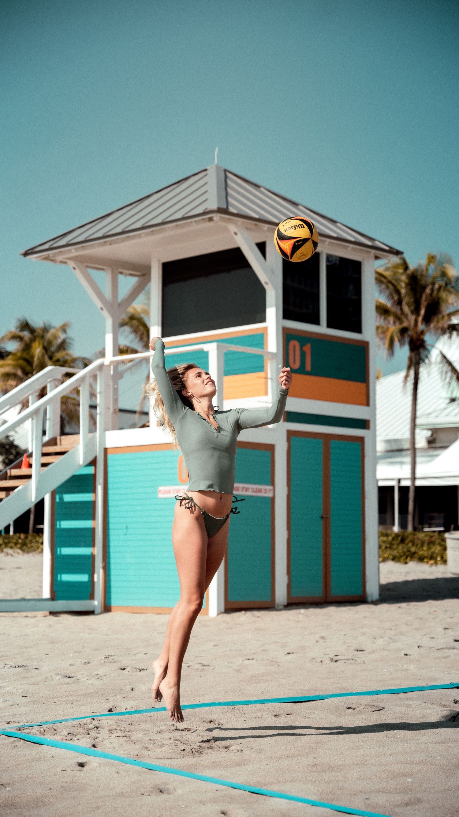 Athletic figure playing beach volleyball near a colorful lifeguard tower in tropical setting.