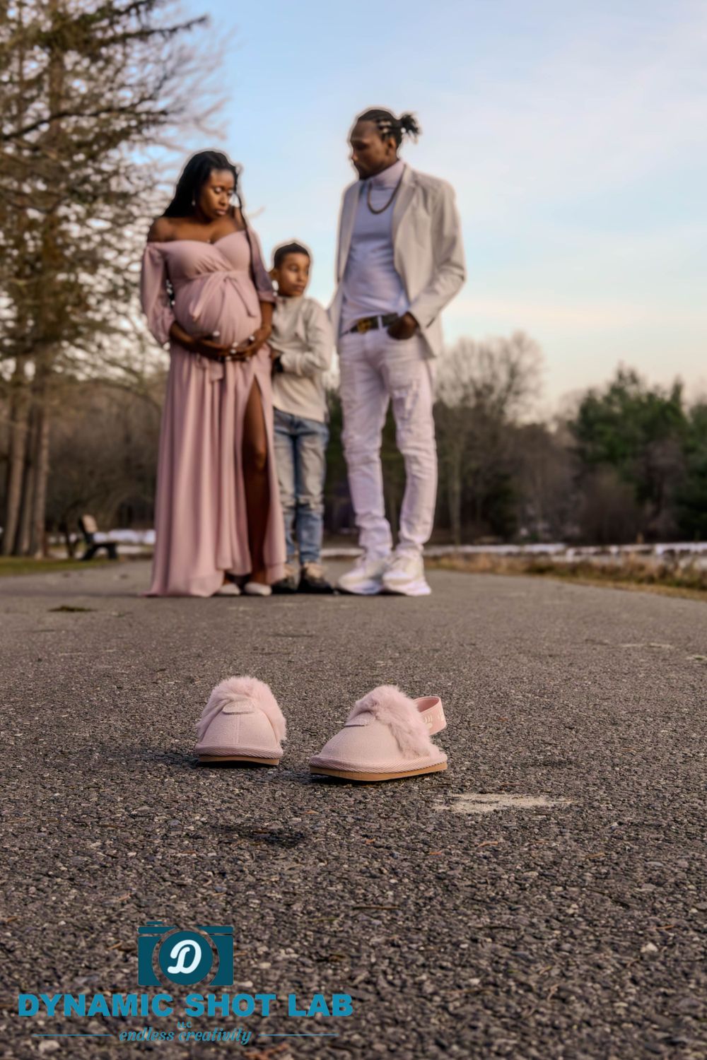 Family maternity photo with pink baby shoes in foreground on asphalt.
