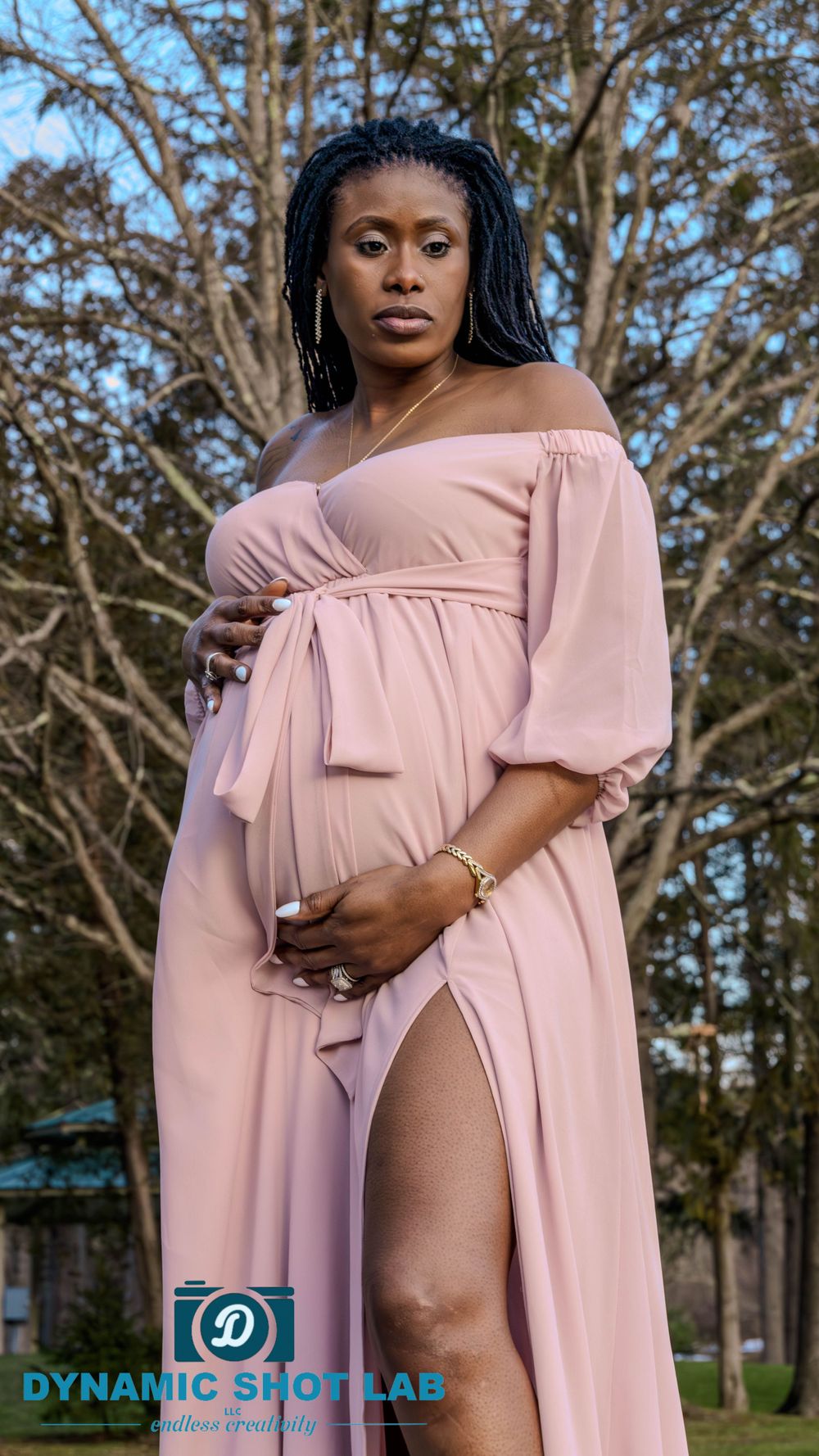Maternity portrait in a flowing pink off-shoulder dress against a natural backdrop.
