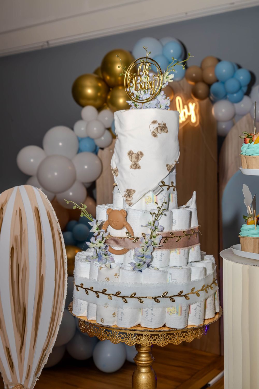 An elegant white and gold tiered diaper cake with teddy bear decorations and balloon garland.