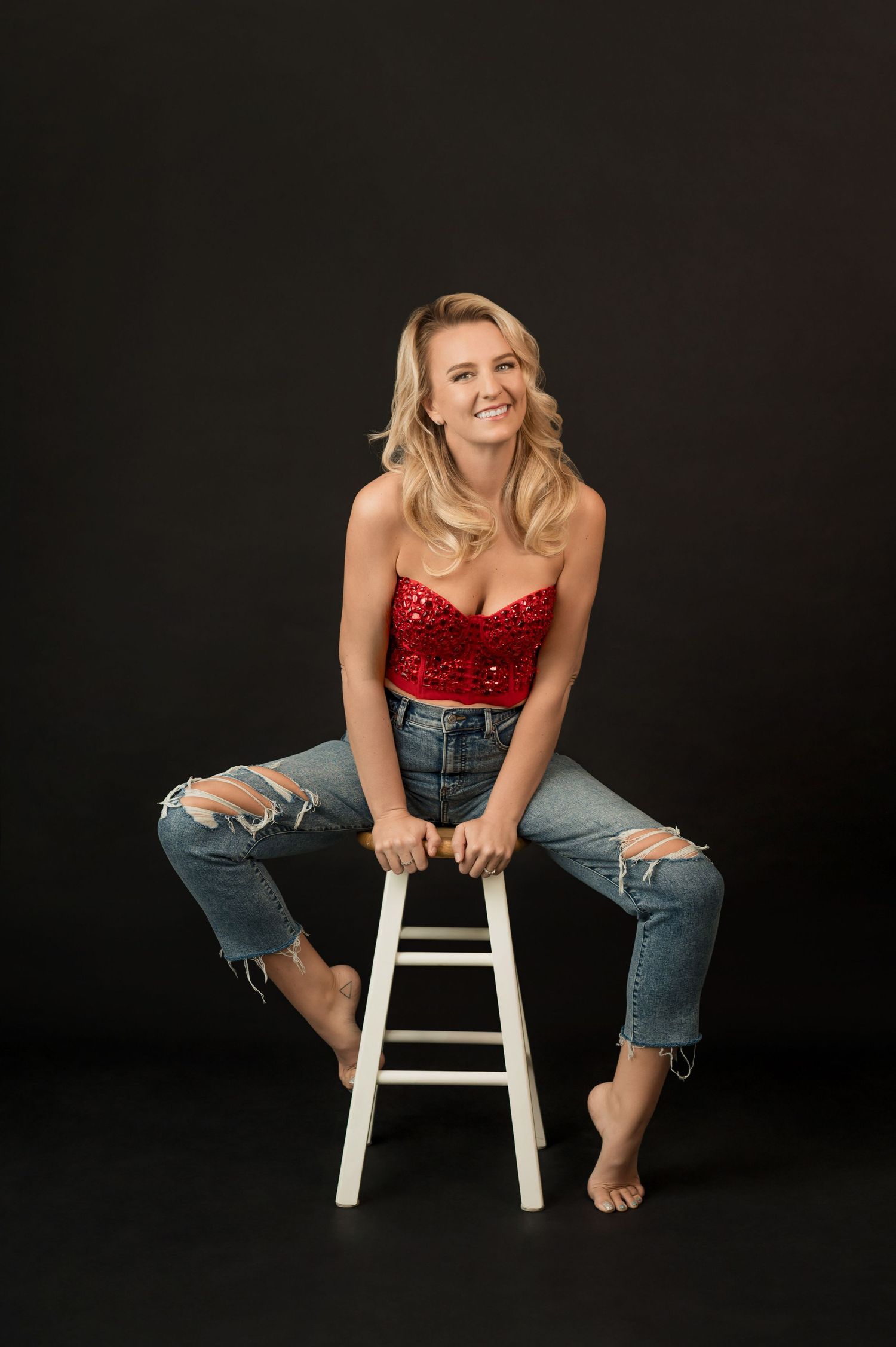 Casual portrait of person in red bustier and ripped jeans sitting on white stool.