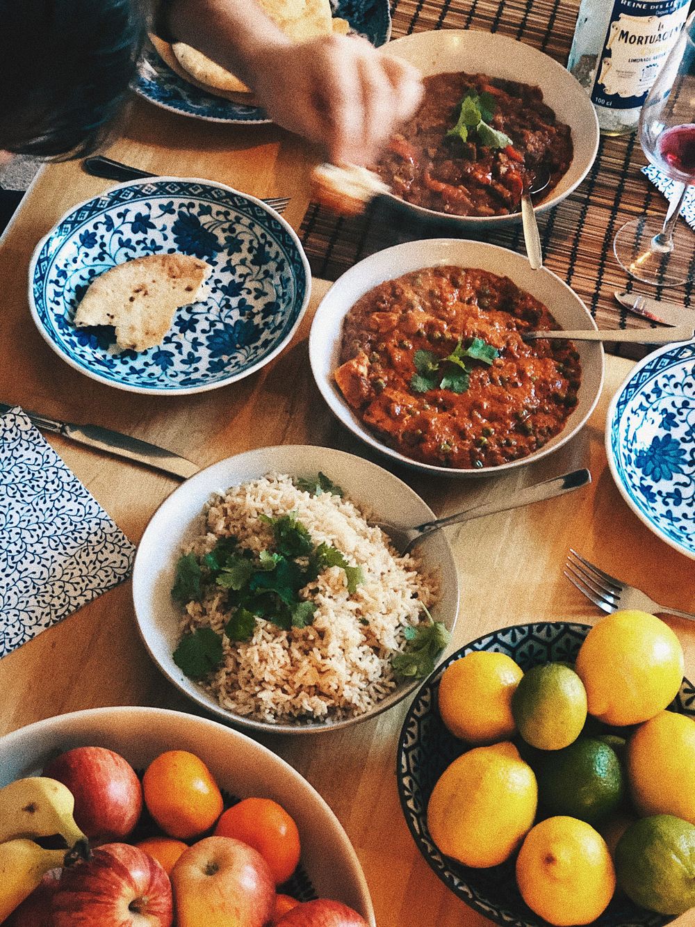 Homestyle curry dishes with rice, fresh fruit, and blue patterned plates on a wooden table.