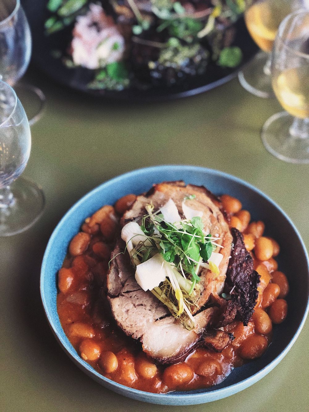 Sliced roasted meat with beans and microgreens in a blue bowl surrounded by wine glasses.