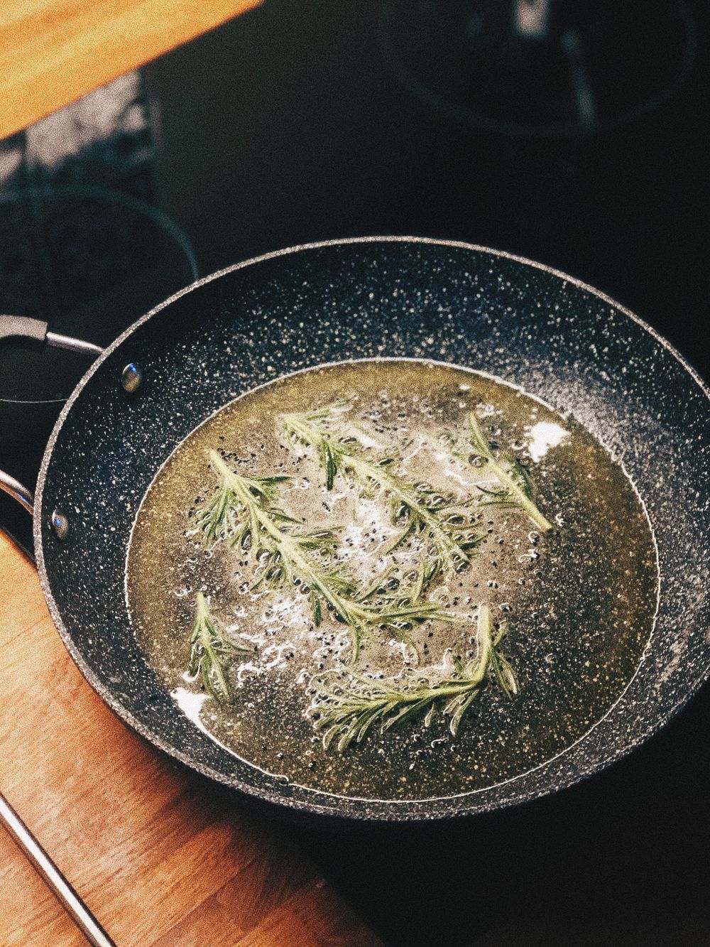 Rosemary sprigs simmering in oil in a black non-stick frying pan.