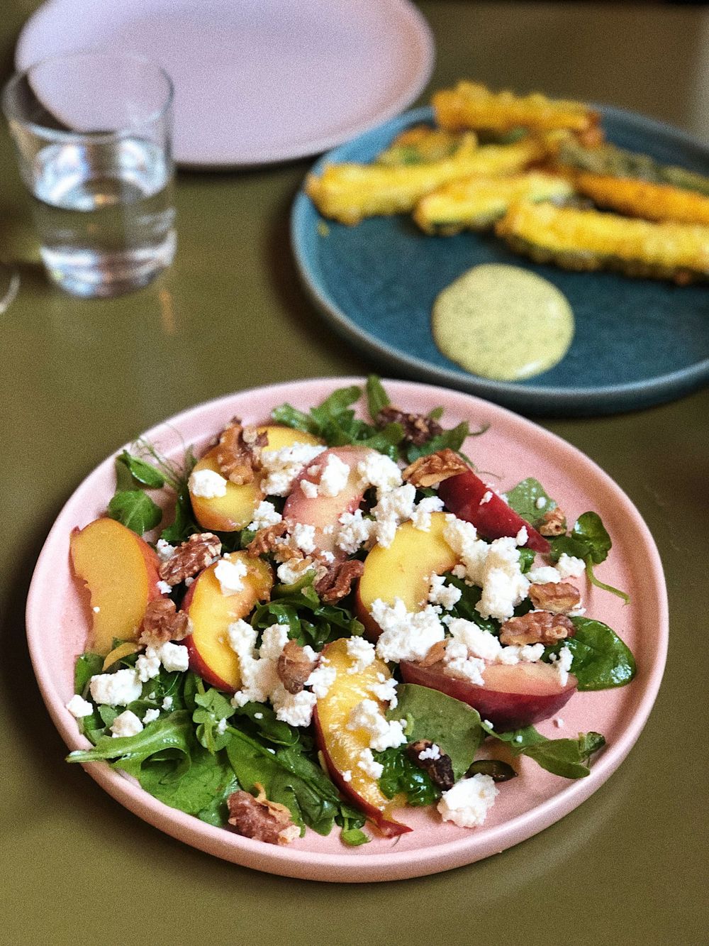 Fresh summer salad with peaches, feta cheese, and nuts on a pink plate next to breaded appetizers.