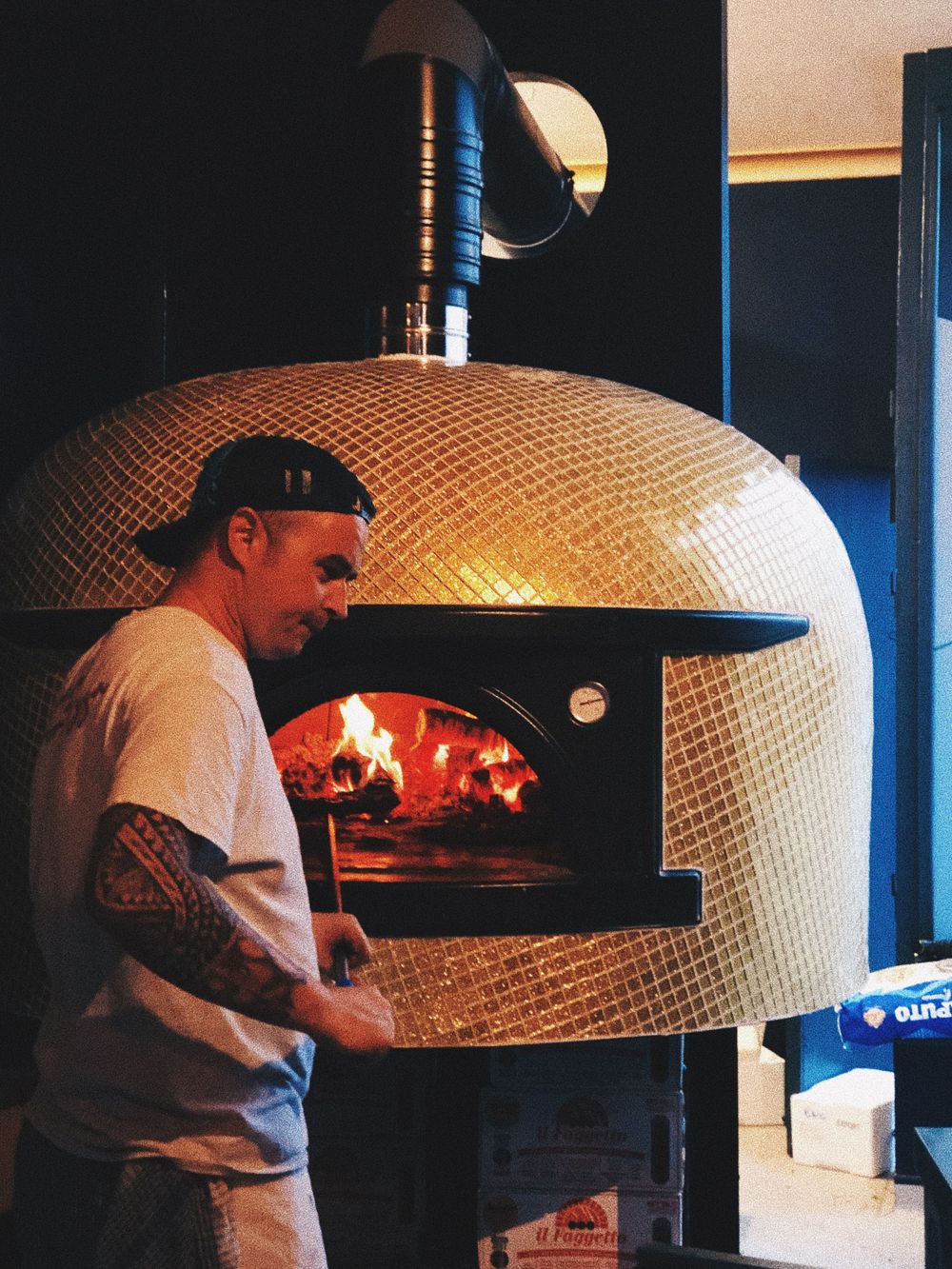 Pizza maker tending to a wood-fired brick oven with flames visible inside.
