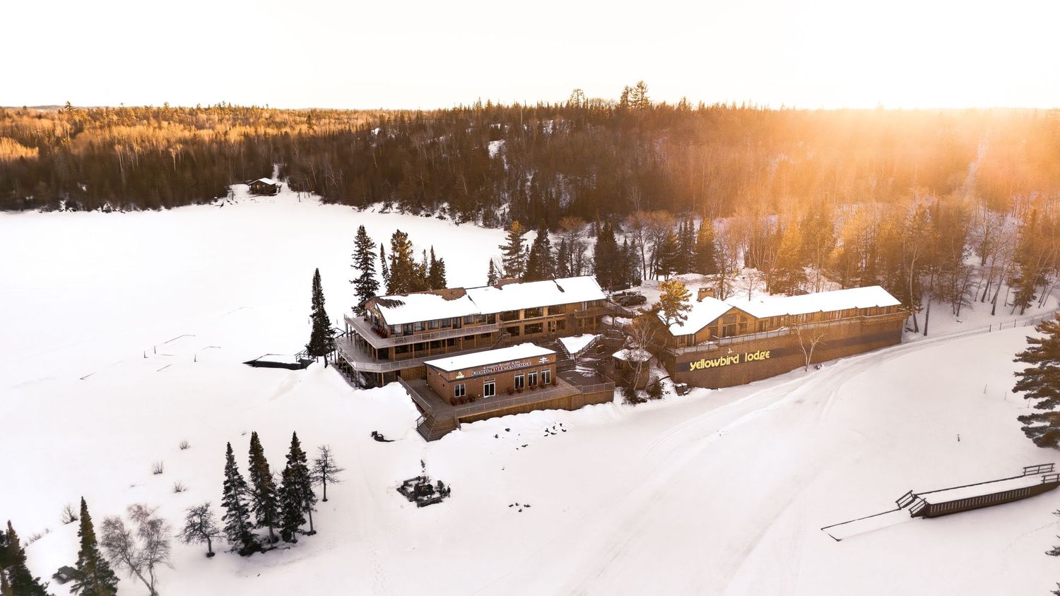 Aerial winter view of a snow-covered lodge surrounded by pine trees and golden sunset light in a remote wilderness setting.