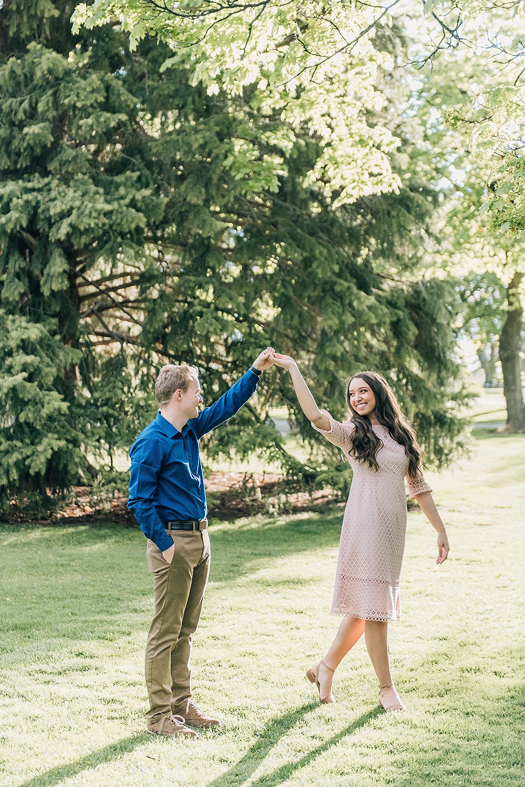A couple dances and twirls together on a grassy lawn surrounded by evergreen trees.