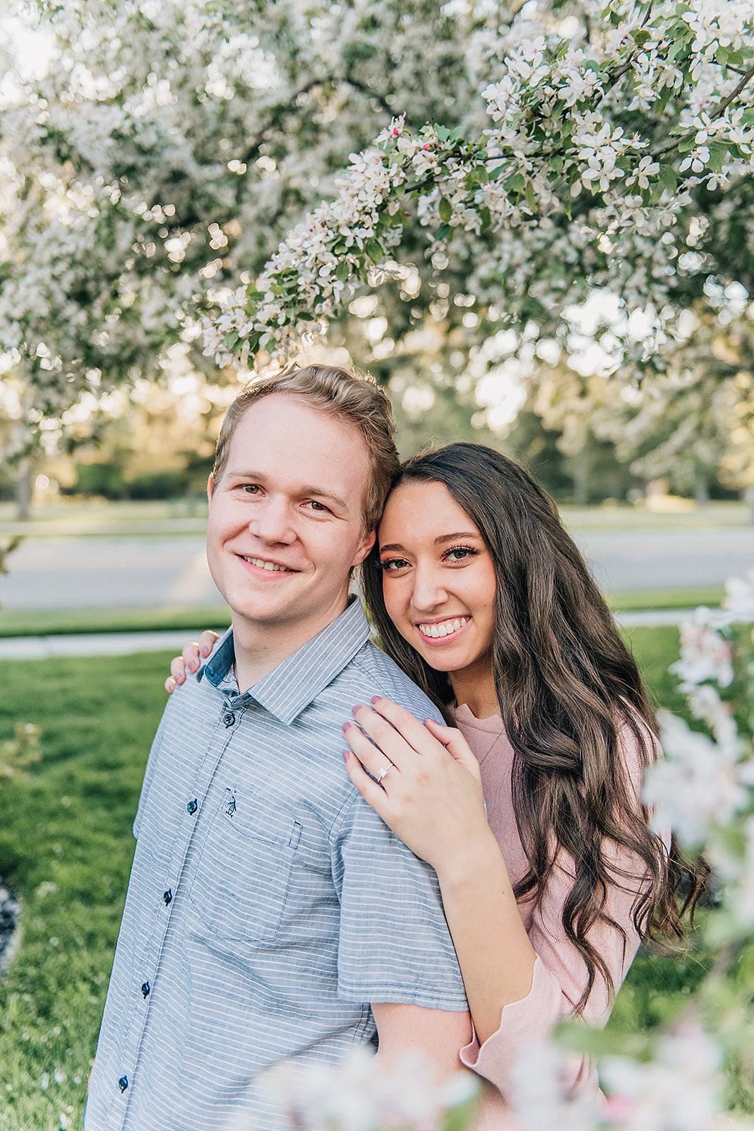 A couple poses among blooming spring cherry blossoms in a park setting.