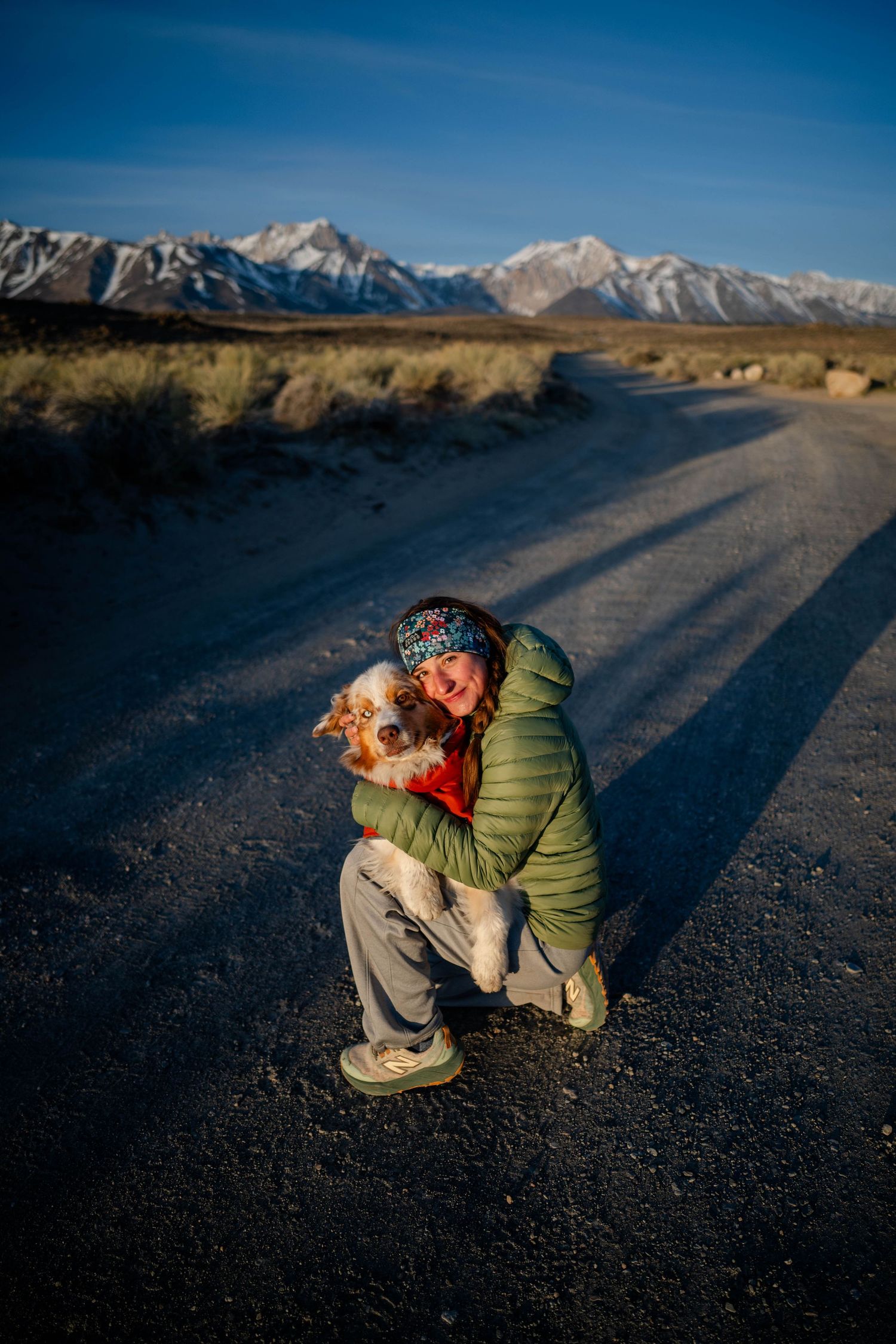 Girl hugging her Australian shepherd crouching on a dirt road with snow-capped mountains in the background.
