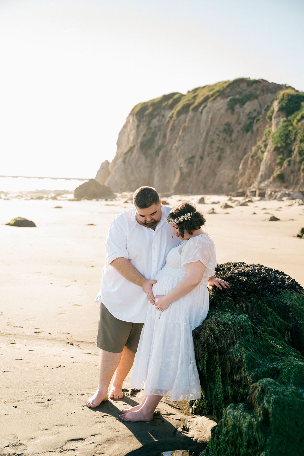 Couple sharing a tender moment on a sandy beach, woman in white dress with floral crown, man in white shirt, rocky cliffs behind.