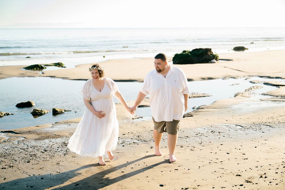 Pregnant couple holding hands walking on a sunny beach, woman in white dress with floral crown, man in white shirt and khaki shorts.