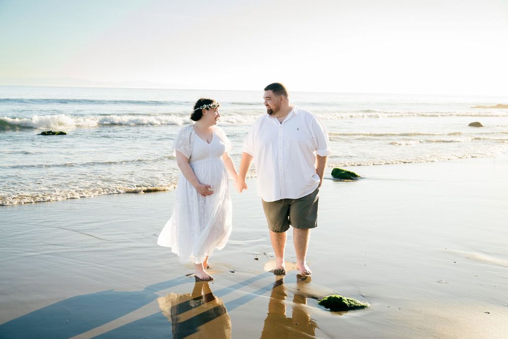 Couple holding hands walking barefoot on a sunny beach, woman in white dress, man in white shirt and shorts.