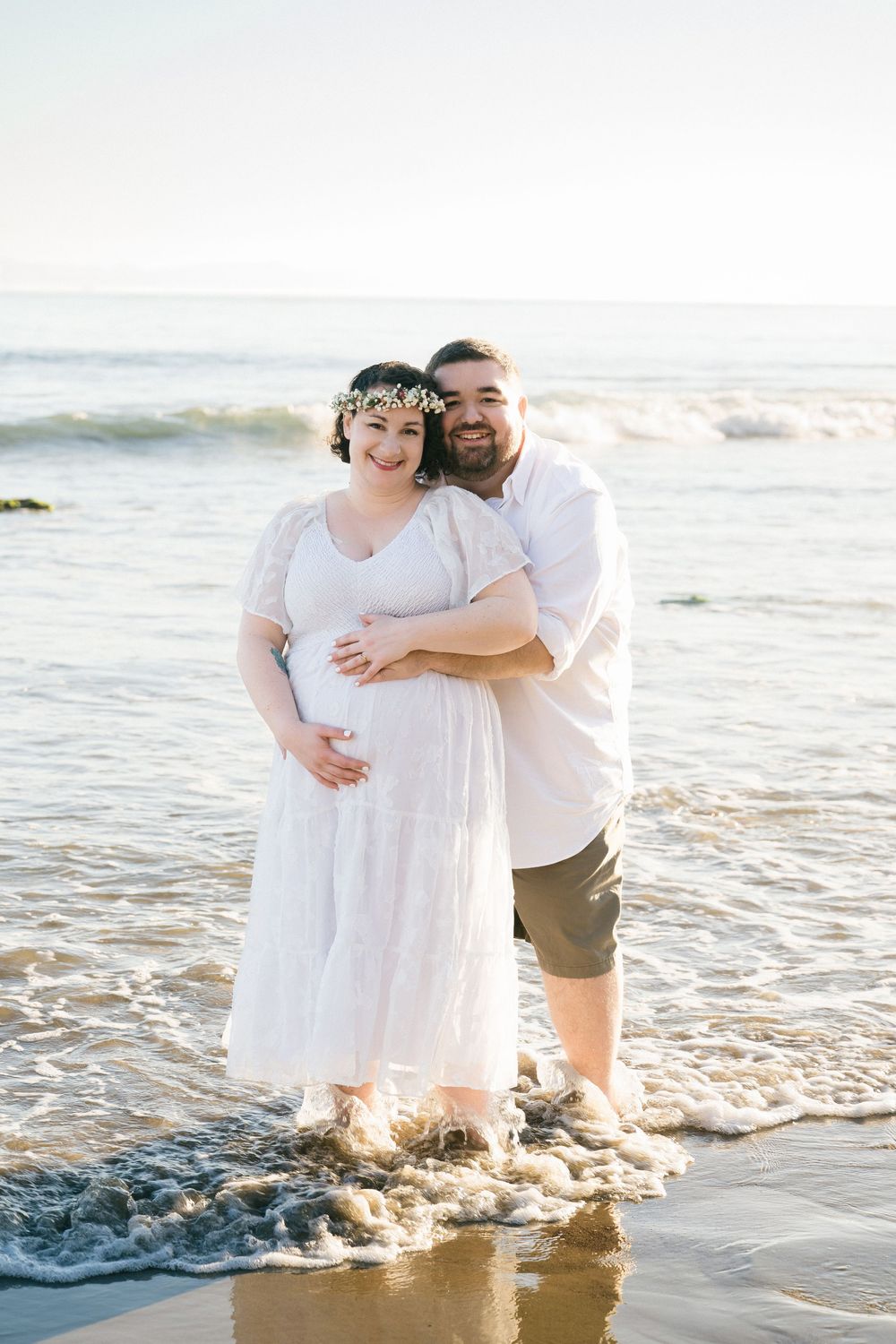 Couple posing at beach shoreline, pregnant woman in white dress with floral crown embraced by man in white shirt, waves at their feet.